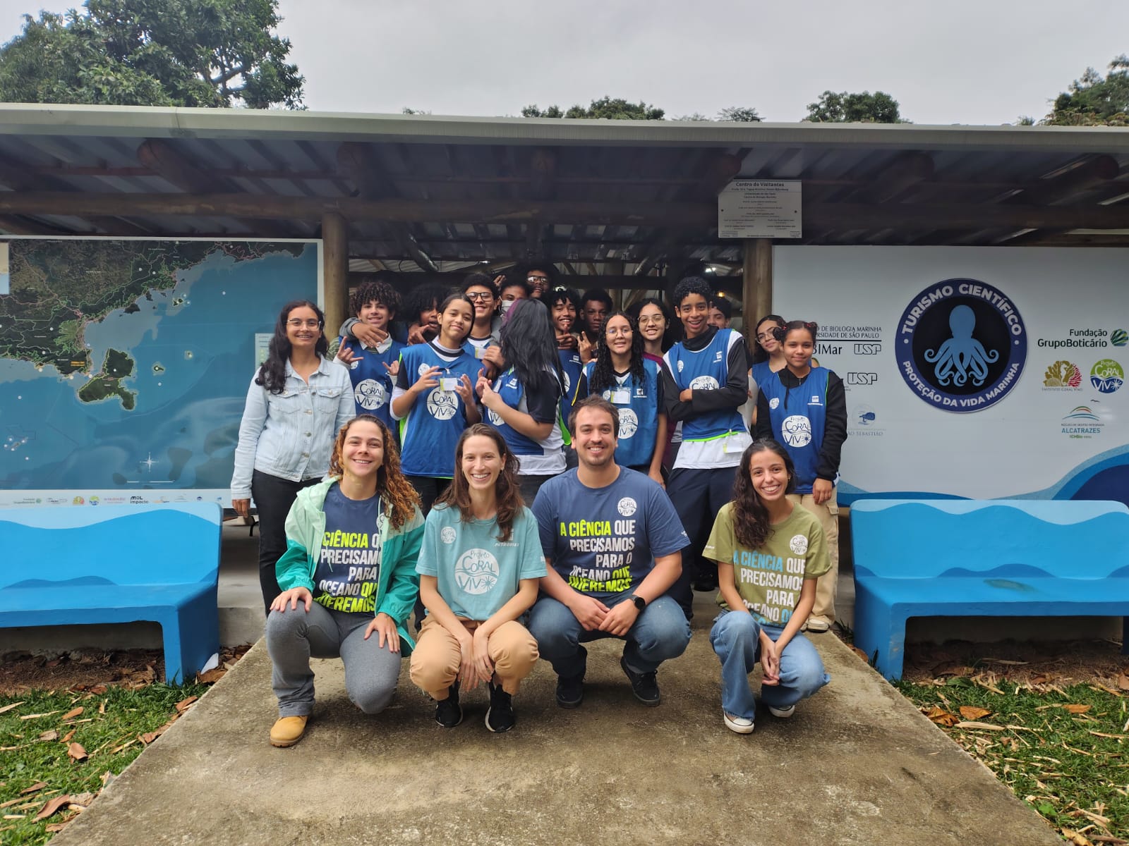 Foto em grupo com cerca de 20 jovens e educadores em frente à estrutura do Centro de Visitantes / Turismo Científico, vestindo coletes azuis e camisetas com a frase "A ciência que precisamos para o oceano que queremos".