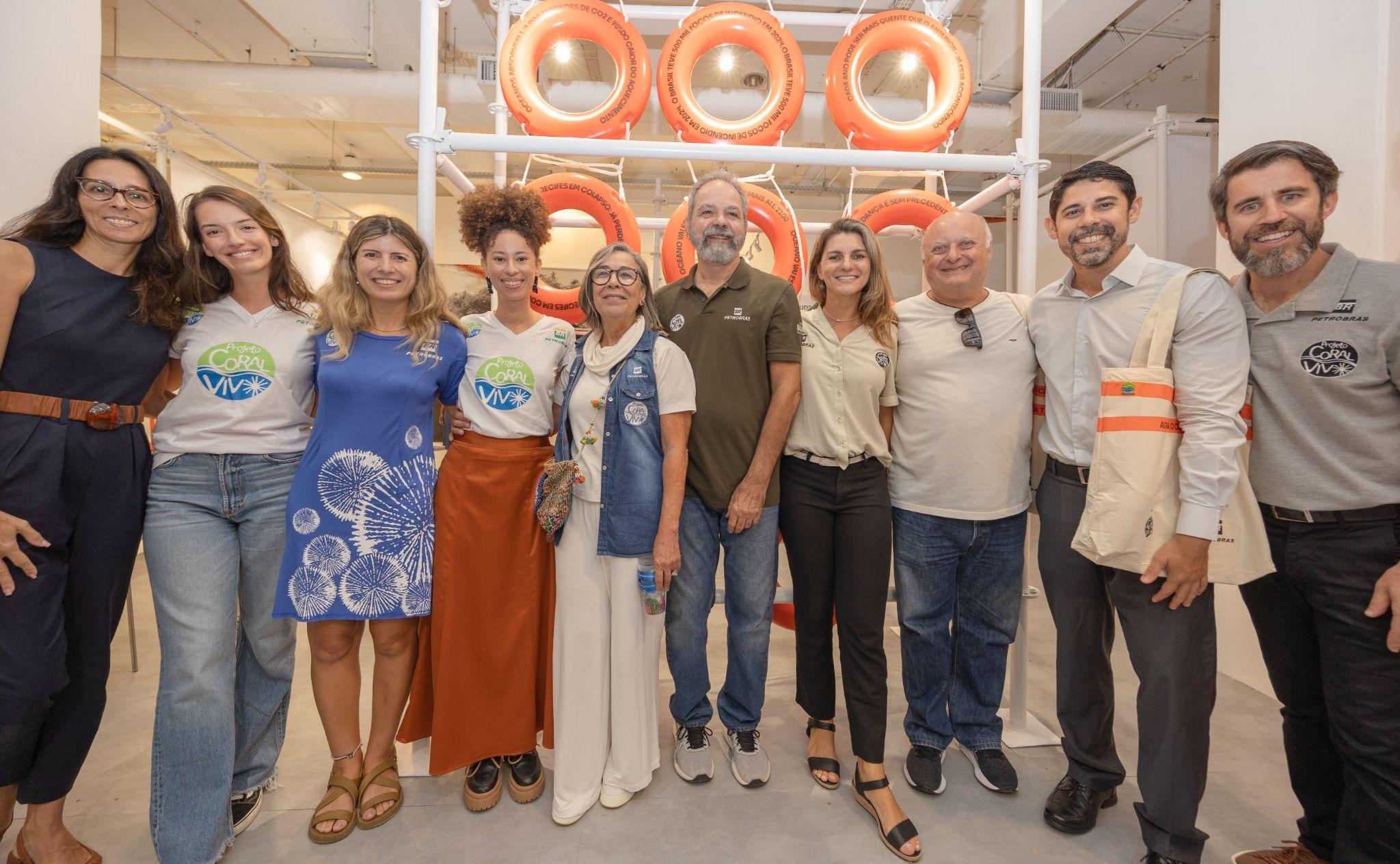 Grupo de homens e mulheres sorrindo lado a lado e posando para foto em frente ao painel temático da exposição sobre clima e oceanos.