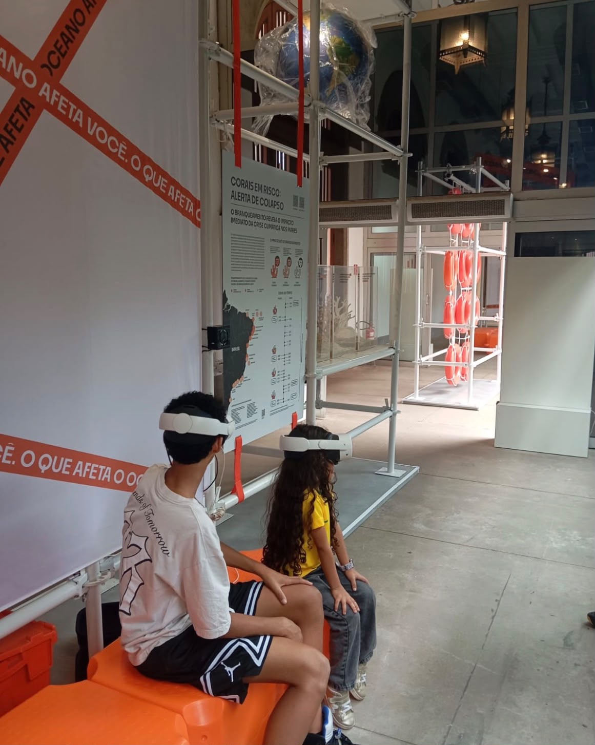 Duas crianças sentadas em bancos de madeira usando óculos brancos de realidade virtual enquanto interagem com a experiência imersiva do oceano.