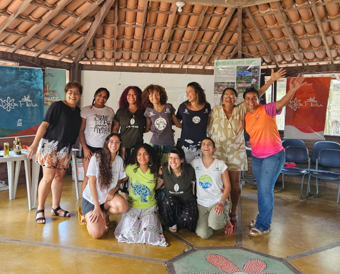 Integrantes do Projeto Coral Vivo e do Coletivo Maré Jovem sorriem reunidos em volta de uma arte de peixe no chão durante o projeto Aiyê Mar.