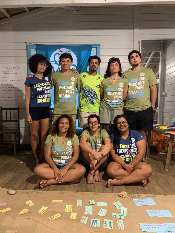 Sete integrantes do Projeto Coral Vivo reunidos em torno de papéis e cartões dispostos no chão durante uma dinâmica de planejamento.
