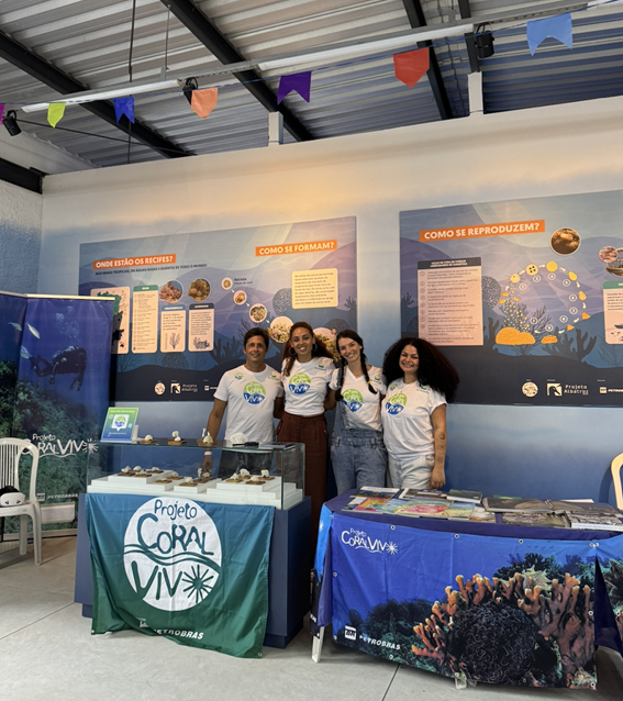 Quatro integrantes da equipe do Projeto Coral Vivo sorriem em um estande de exposição com mesas e painéis educativos sobre recifes de corais.