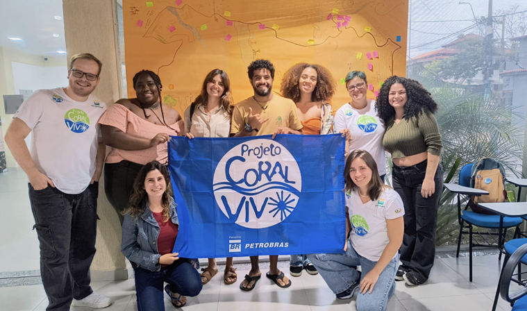 Seis integrantes da equipe do Projeto Coral Vivo sorriem segurando uma grande bandeira azul do projeto em um ambiente interno.